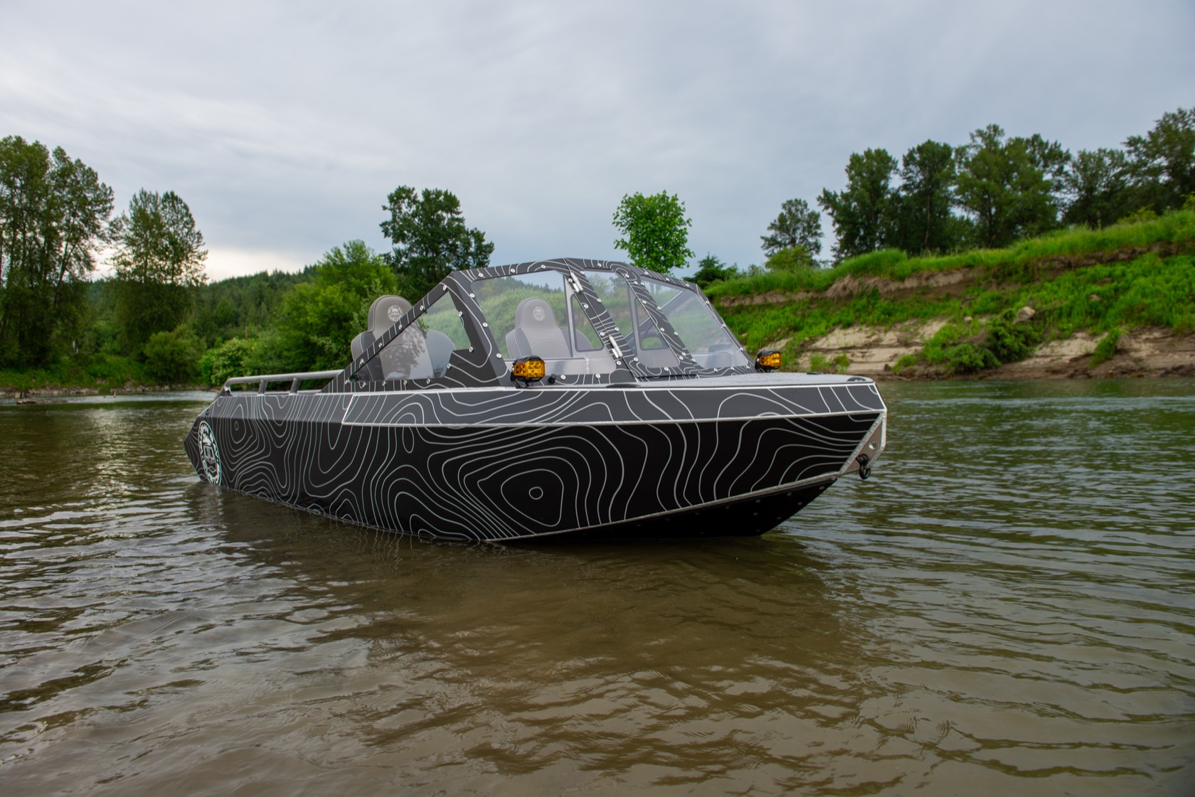 Front-quarter view of the topo-wrap jet boat floating on the green river with a forested bank behind