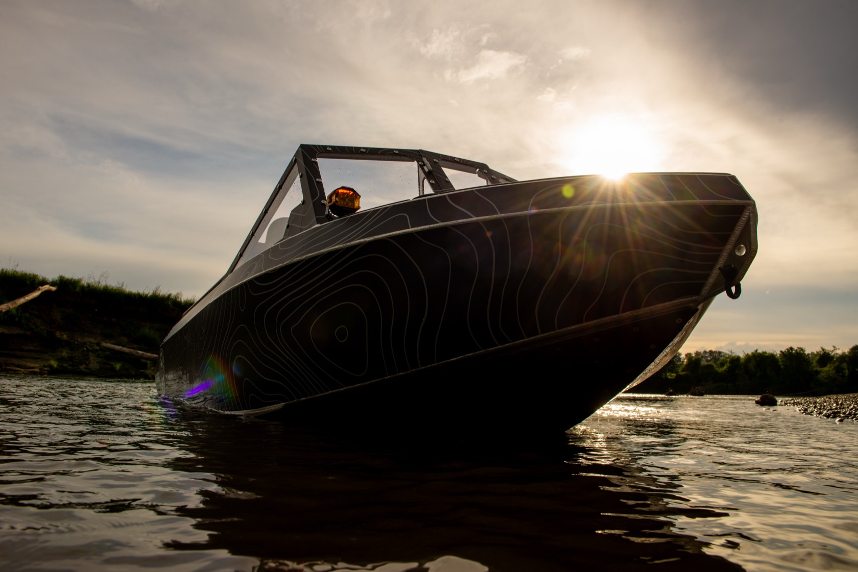 Low front-quarter view of the black topographic-wrap Vaughn Bay Customs jet boat at rest on the river, the sun bursting over its bow