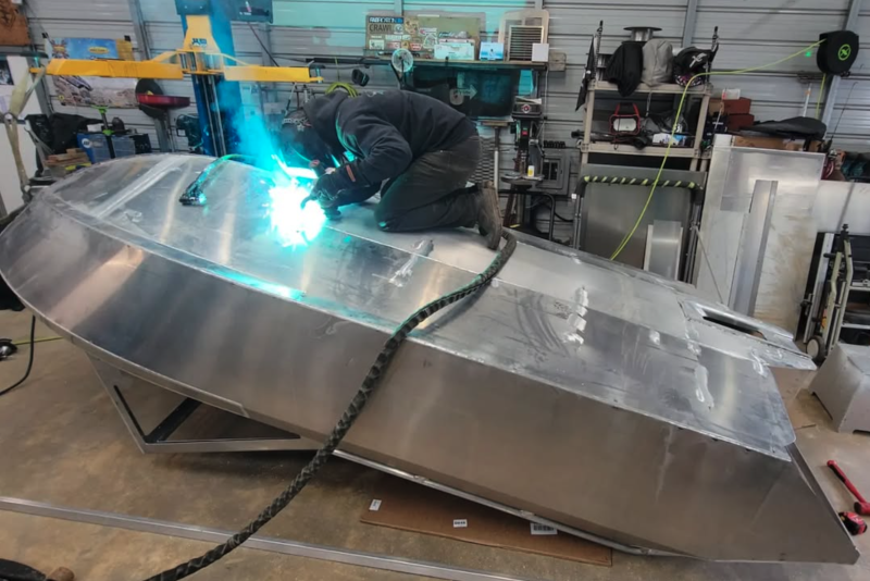 Welder joining the top deck seam of a bare welded aluminum jet boat hull under a bright arc