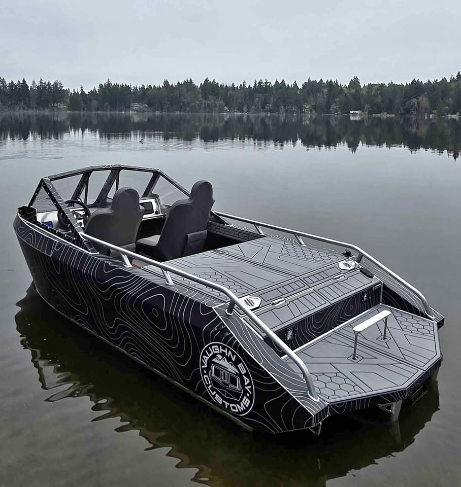Rear-quarter view of the Vaughn Bay Customs VB12 show boat on a calm lake — black topographic-wrap hull with the circular VBC mountain logo