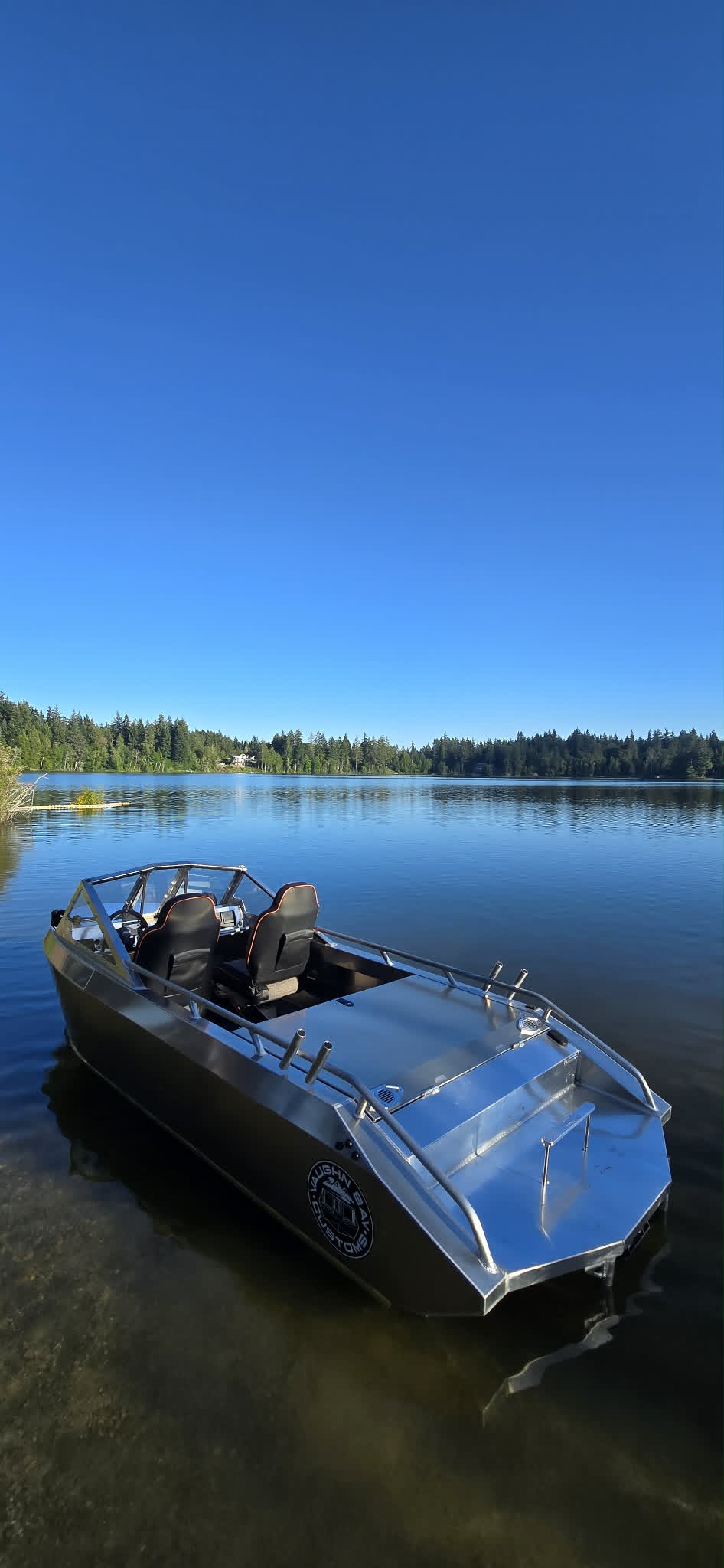 Front-quarter view of a Vaughn Bay Customs VB14 welded aluminum jet boat floating on a calm forest lake, black hull and raw aluminum deck